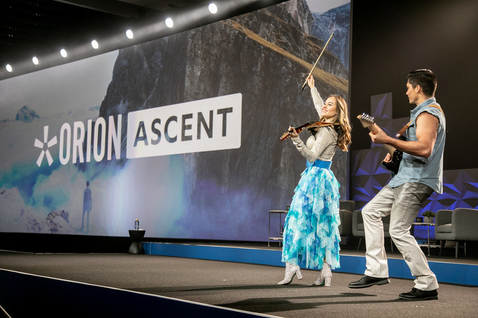 Photo of 2 musicians, a violinist and a guitarist, performing on stage in front of LED screen showing the Orion Ascent logo