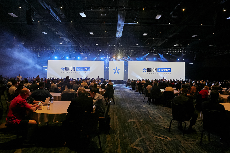 Photo of around 200 audience members sitting at round tables and looking at a stage with Orion Ascent Conference 2023 backdrops and blue lighting