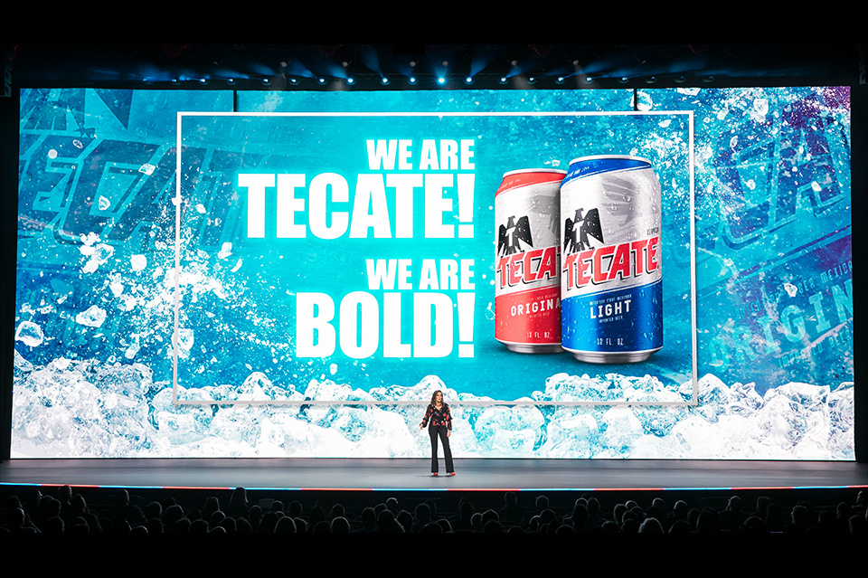 Photo of a presenter on stage in front of a blue LED background showing Tecate cans with the text "We are Tecate, we are bold!" Prester is addressing a large audience
