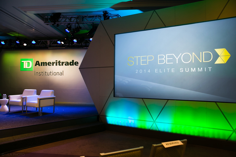 Photo of an empty TD Ameritrade Institutional stage with green lighting. Twp empty white chairs are set on stage next to a screen tat reads "Step Beyond 2014 Elite Summit"
