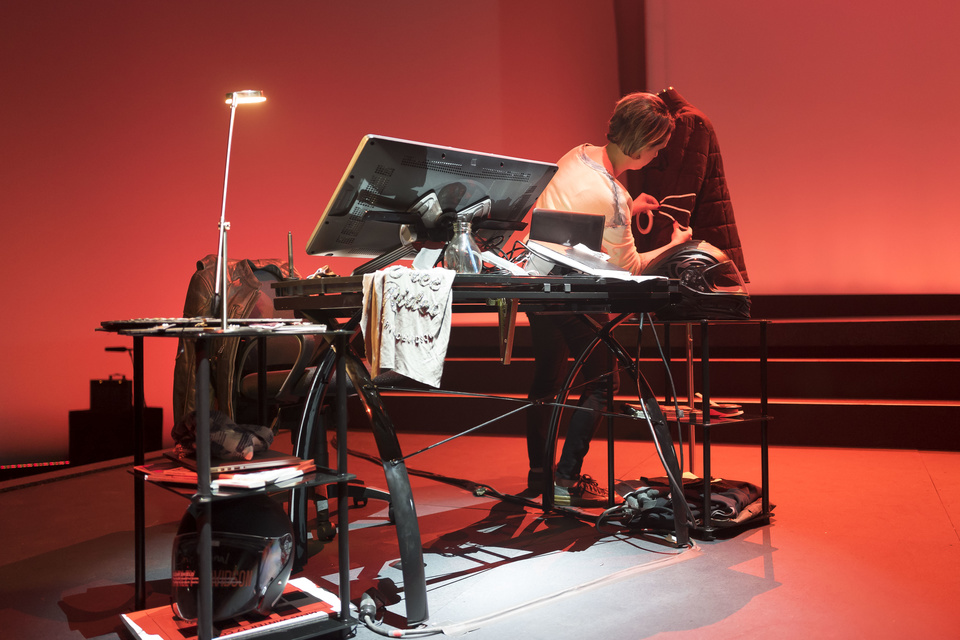 Photo of woman measuring the sleeve of a jacket next to a work desk. Set is on stage.