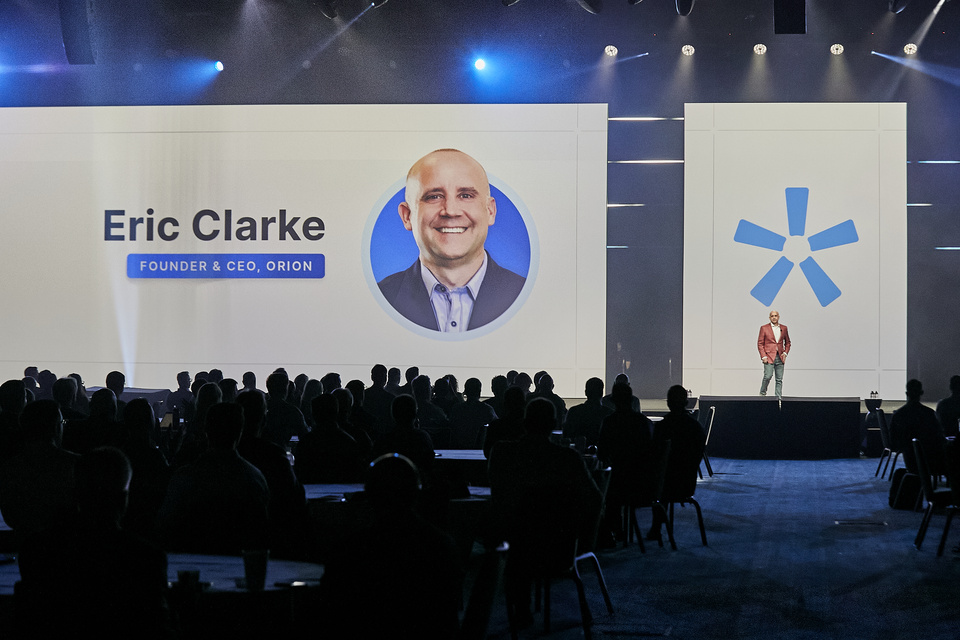 Photo of a presenter, Eric Clarke, Founder & CEO, Orion speaking to an audience in front of a title slide with his headshot on an LED screen