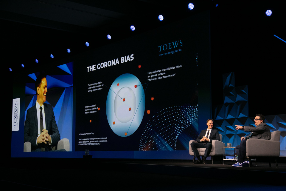 Image of 2 presenters sitting in chairs on stage presenting information in front of an LED screen reading "The Corona Bias"