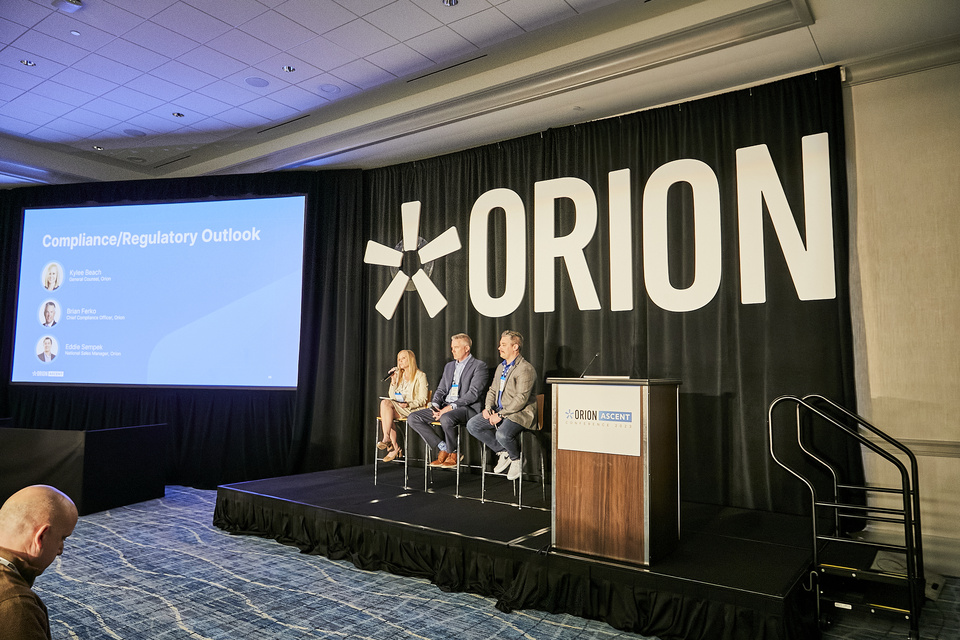 Photo of 3 presenters on a small stage with an Orion logo backdrop. A screen to the left of the speakers reads "Compliance/Regulatory outlook.