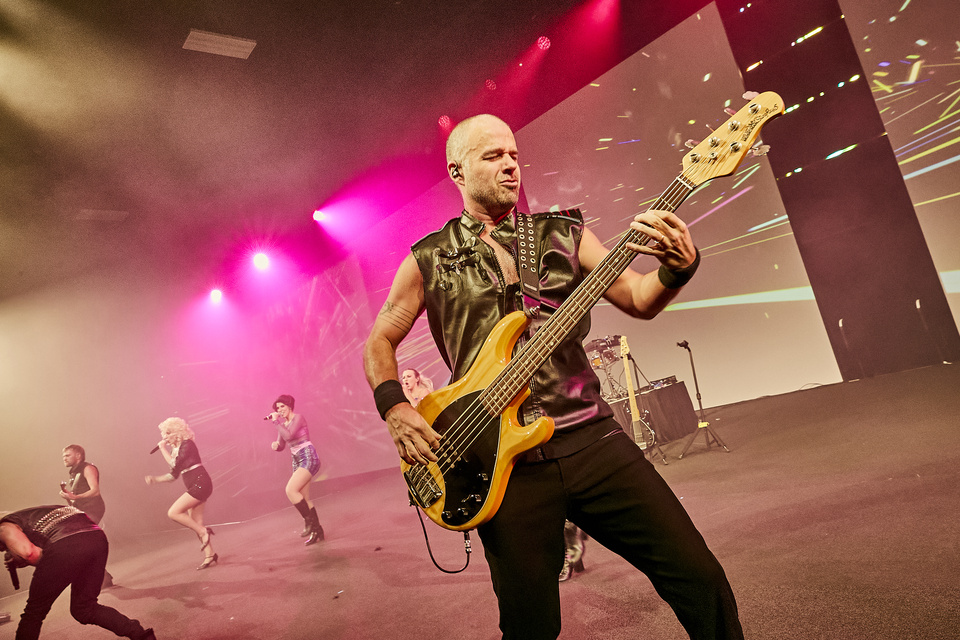 Photo of a rock musician playing a bass guitar on a stage with other band members, singers and guitarists, playing in the background