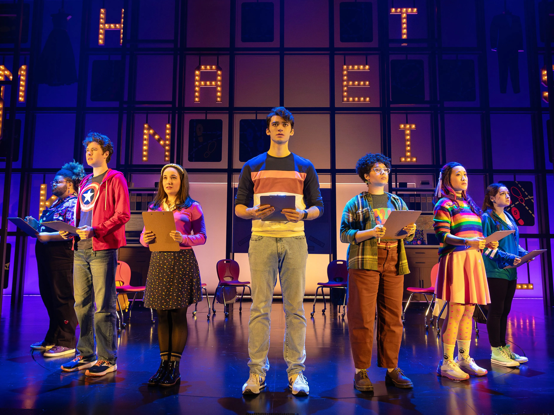 Image of 7 actors on a stage holding clipboards in front of a classroom set