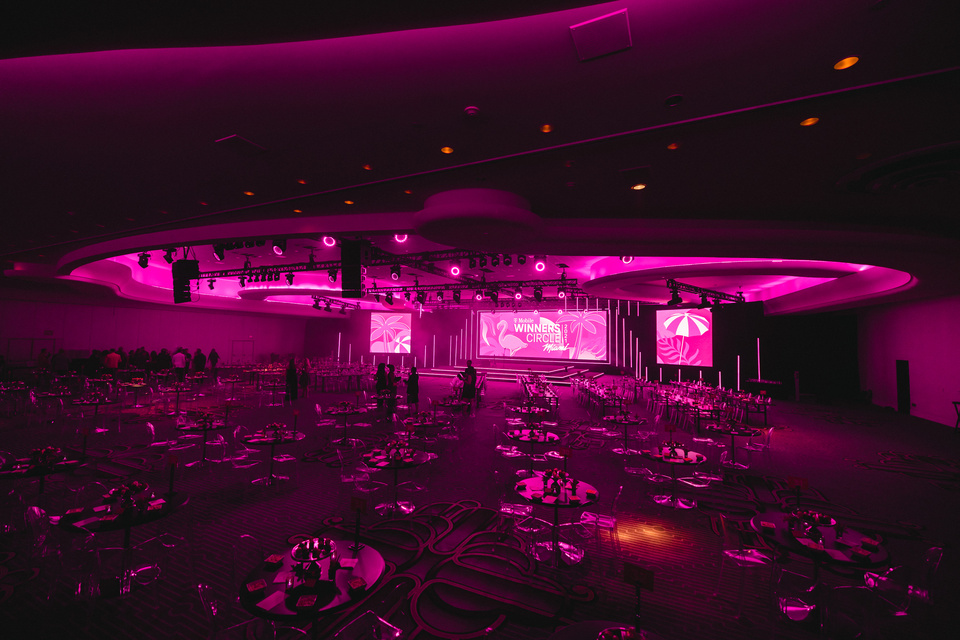 Large event room setup with magenta lighting, round tables, and a stage displaying 'T-Mobile Winner's Circle' with tropical-themed graphics.