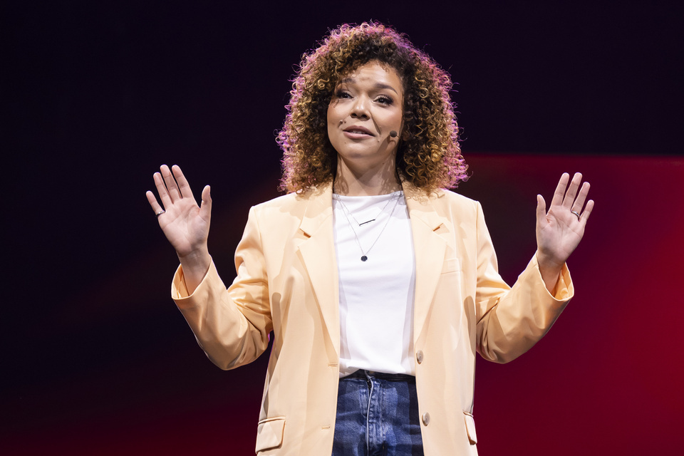 Photo of a woman in a yellow blazer speaking into a microphone with both hands held up.