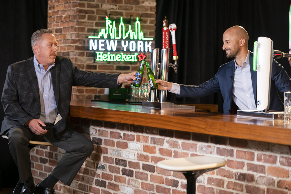 Photo of Two Men cheersing Heineken bottles at staged bar in front of a New York Heineken Neon Sign