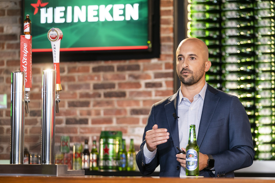 Photo of a Man speaking from behind a staged bar with a Heineken logo on TV behind him