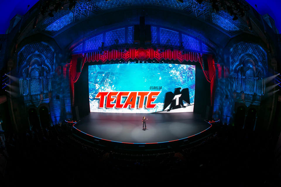 Photo of presenter on stage in a theater in front of an LED screen showing Tecate logo