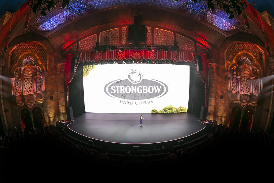 Photo of presenter on stage in a theater in front of an LED screen showing Strongbow Hard Ciders logo