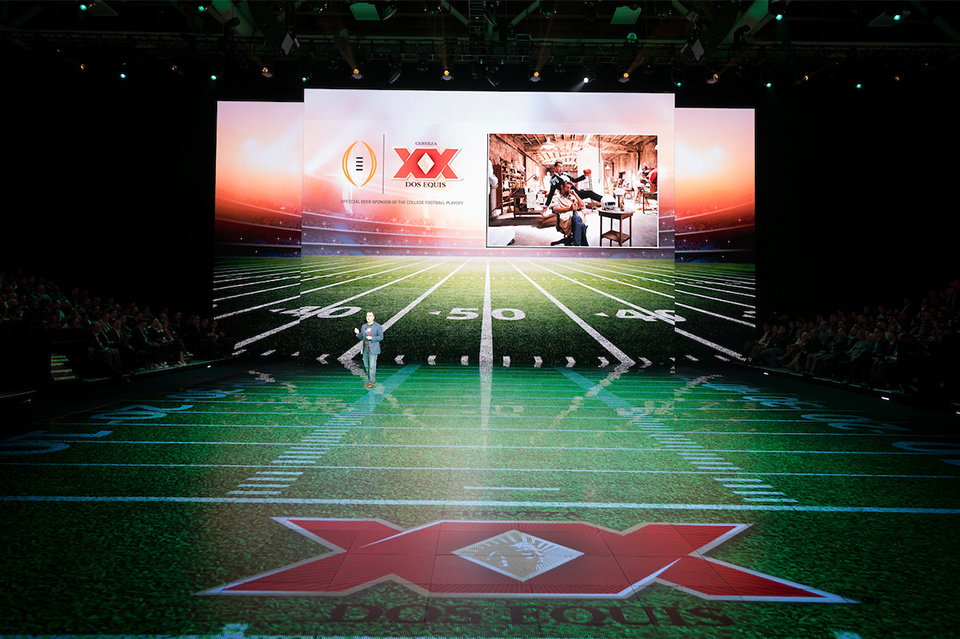 Photo of presenter on XX stage. LED screens show a football field set.