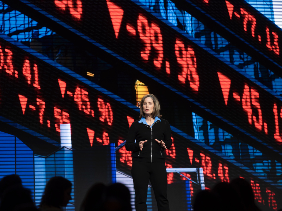 Image of a woman on stage speaking with a stock ticker backdrop