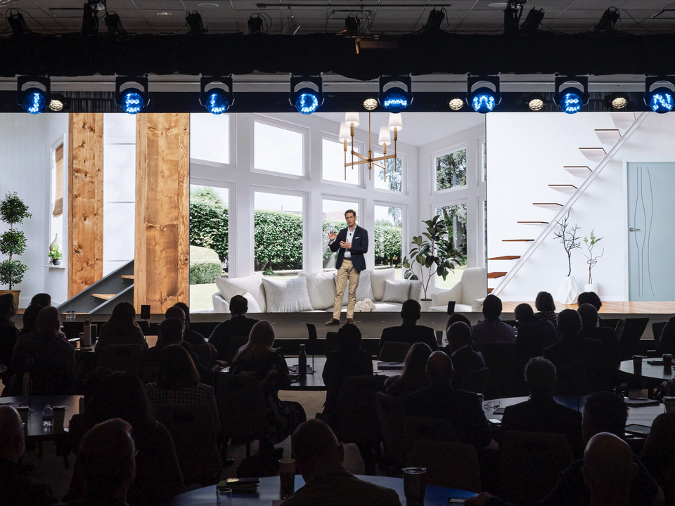 image of a presenter on stage speaking to a group of people sitting at round tables in front of a realistic clean home background and blue custom JELD-WEN lights