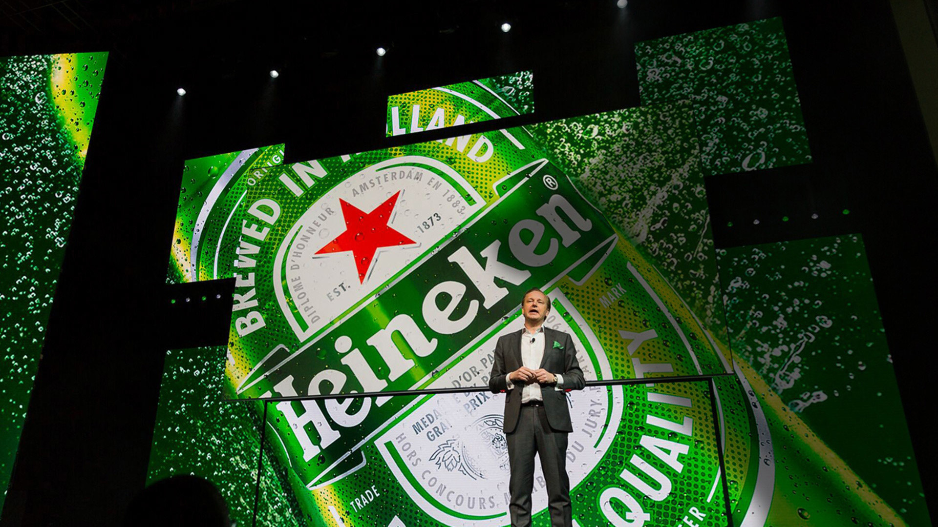 Photo of 1 presenter on stage in front of an LED screen showing a Heineken bottle.