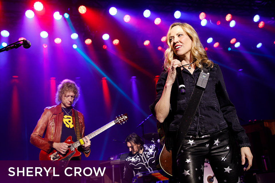 Photo of Sheryl Crow singing into a microphone on stage in front of a band. Bottom left plum footer reads "Sheryl Crow"