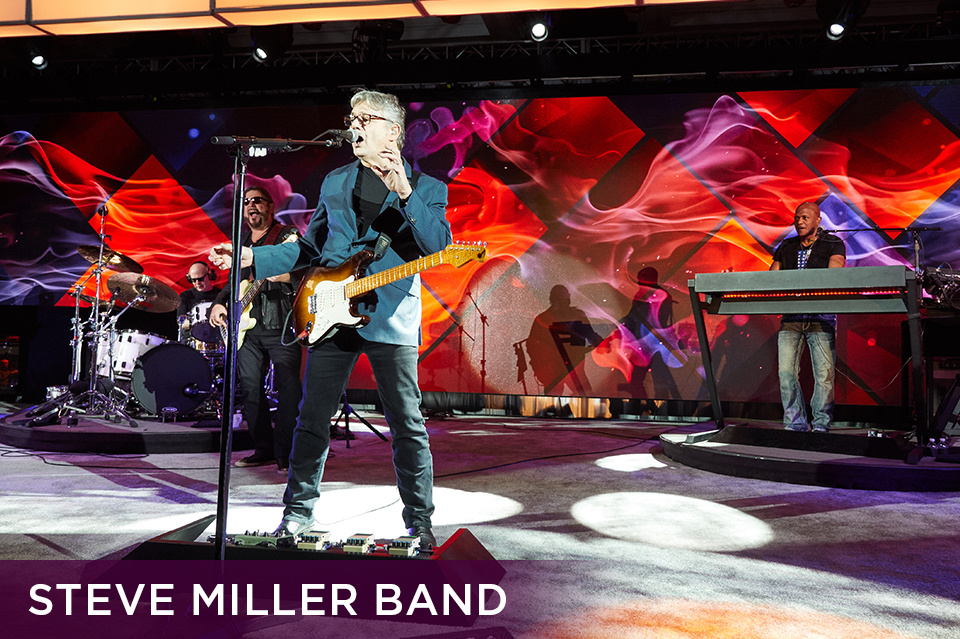 Photo of guitarist of the band Steve Miller Band with an electric guitar singing into a microphone on stage in front of a band. Bottom left plum footer reads "Steve Miller Band"