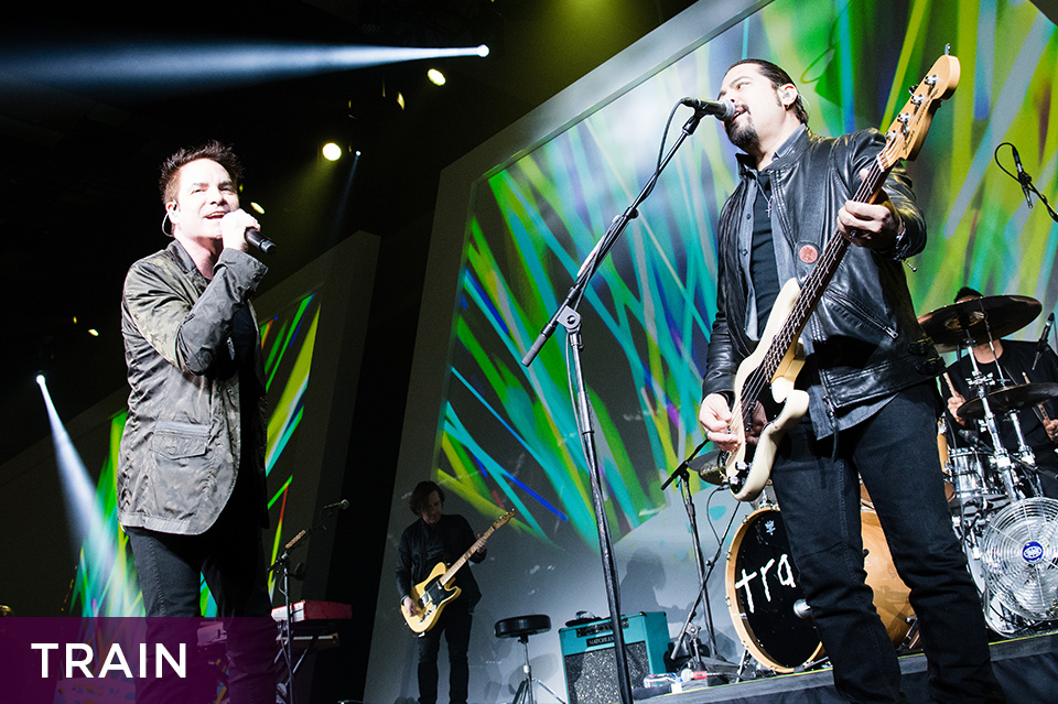 Photo of members of the band Train performing on stage in front of a green design on an LED screen. Bottom left plum footer reads "Train"