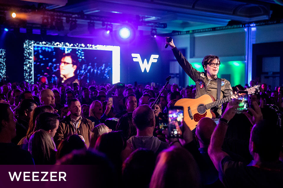 Photo of guitarist of the band Weezer with an acoustic guitar putting a microphone up in the air in the center of a large crowd. Bottom left plum footer reads "Weezer"