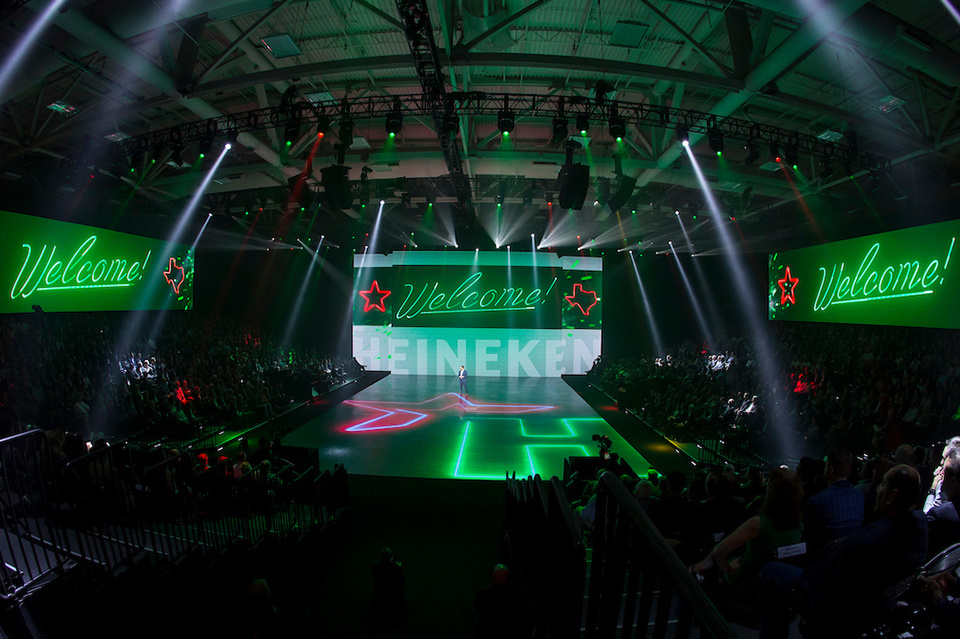 Photo of presenter welcoming an audience on stage in front of an LED screen showing Heineken logo and "welcome!" on 3 LED screens.