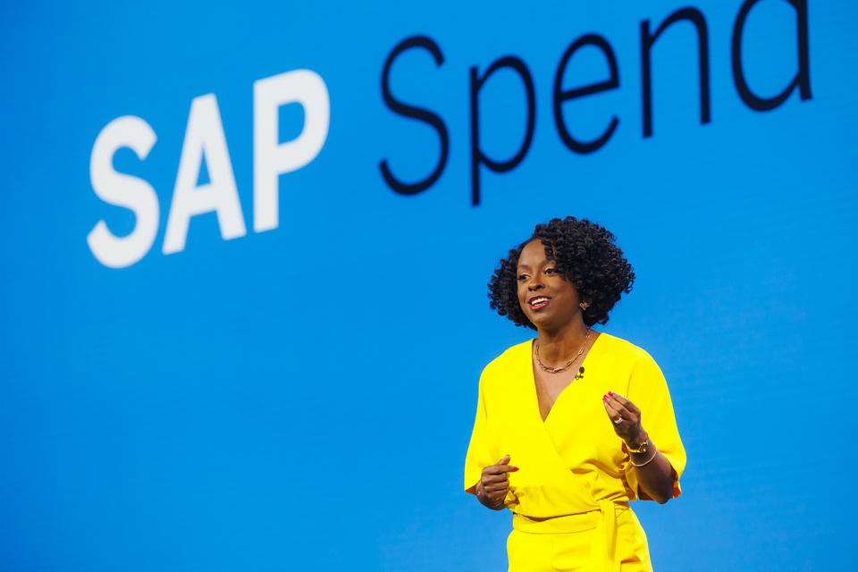 Photo of a woman giving a presentation in front of a blue screen that reads "SAP Spend Connect Live"
