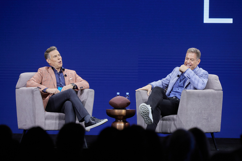 Photo of 2 speakers having a conversation on stage and sitting in grey chairs. A table is between them with a football on it