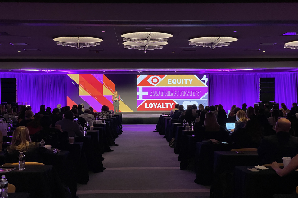 Photo of a presenter on a medium sized stage speaking to an audience in front of a screen that reads "Equity, Authenticity, Loyalty."