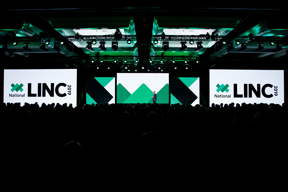 Photo of a man speaking to a large audience on a stage in front of 5 LED screens. 2 screens read National LINC 2019.