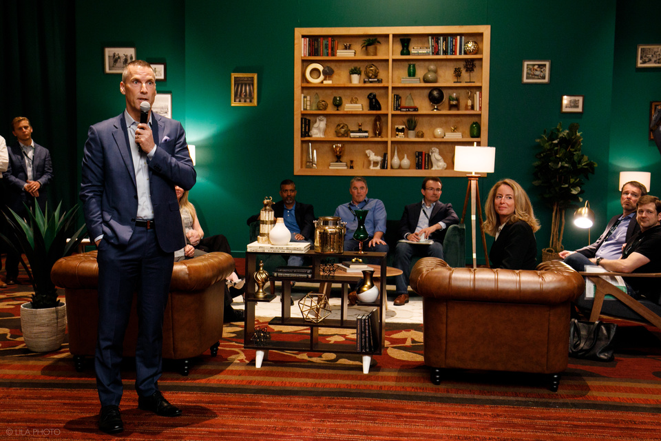 Photo of 10 people in A Modern Living Room Setting. One man is standing in the foreground speaking into a microphone while the other people in the scene look on.