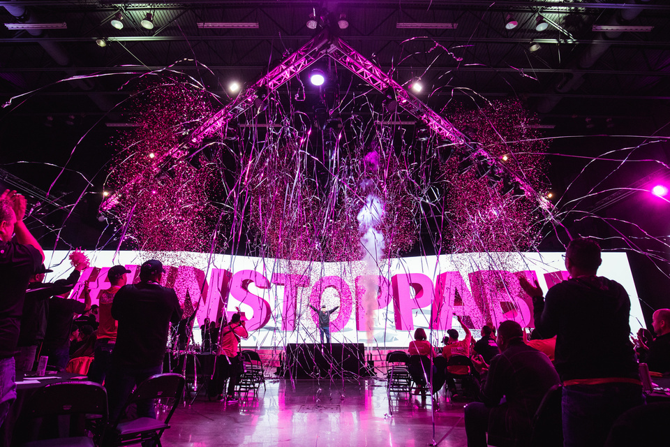Photo of a performer on a platform in front of an LED screen that reads "Unstoppable." Magenta confetti is shot for a small audience.