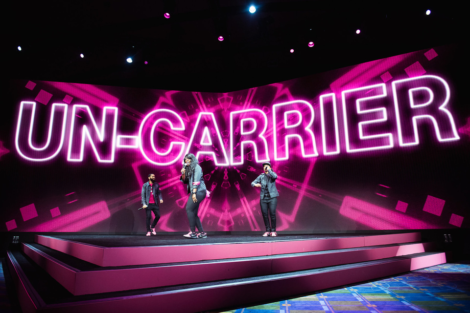 Photo of 3 performers on a magenta stage in front of an LED screen that reads "Un-Carrier"