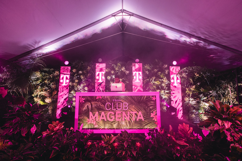 Photo of a tropical stage and DJ booth with T-Mobile Club Magenta branding. DJ is at booth with a laptop.
