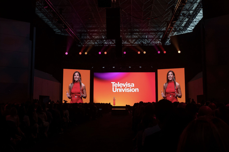 Photo of a woman speaking to an audience on a stage with a Televisa Univision Orange backdrop, wide shot