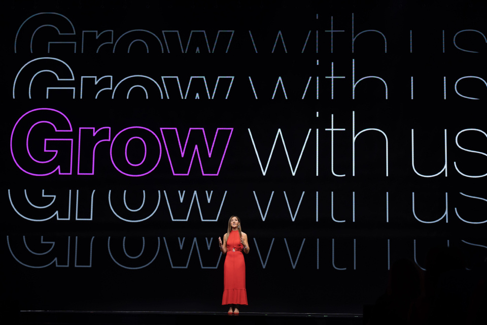 Photo of woman speaking on stage in front of screen that reads "Grow with Us"