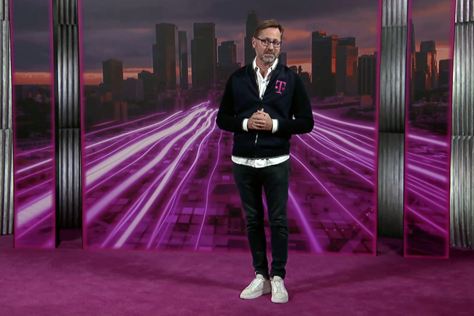 Photo of T-Mobile CEO speaking on a stage with a magenta carpet in front of city backdrop with magenta waves