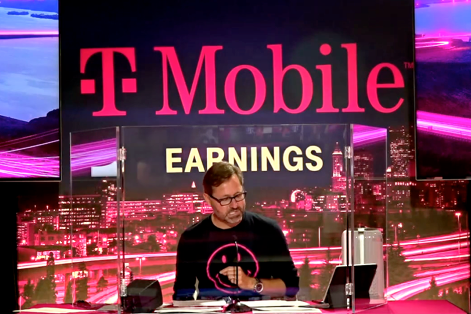 Photo of T-Mobile CEO speaking into a microphone at a desk and looking at an iPad. Backdrop reads "T-Mobile Earnings" in front of city backdrop with magenta waves