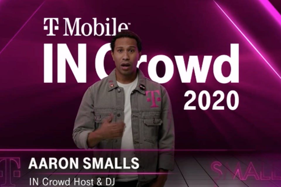 Photo of DJ Aaron Smalls speaking into a microphone behind a DJ booth in front of black backdrop with multiple "T" T-Mobile logos
