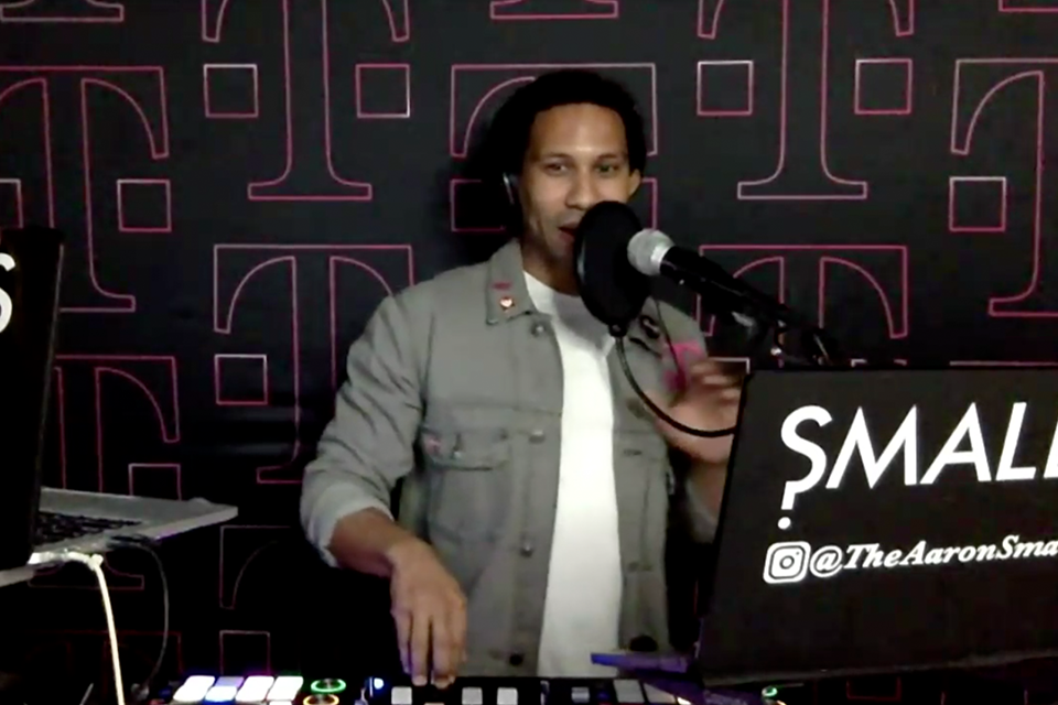 Photo of DJ Aaron Smalls speaking into a microphone behind a DJ booth in front of black backdrop with multiple "T" T-Mobile logos