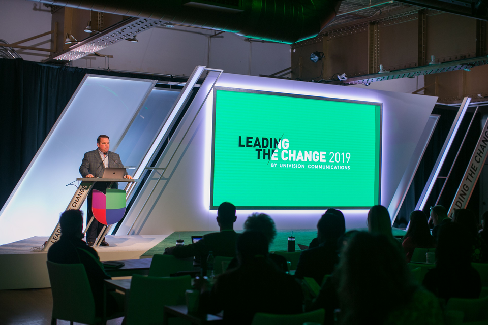 Photo of a speaker at a lectern with a laptop on it presenting to a small crowd. A screen text to the presenter reads "Leading the Change 2019 by Univision Communications."
