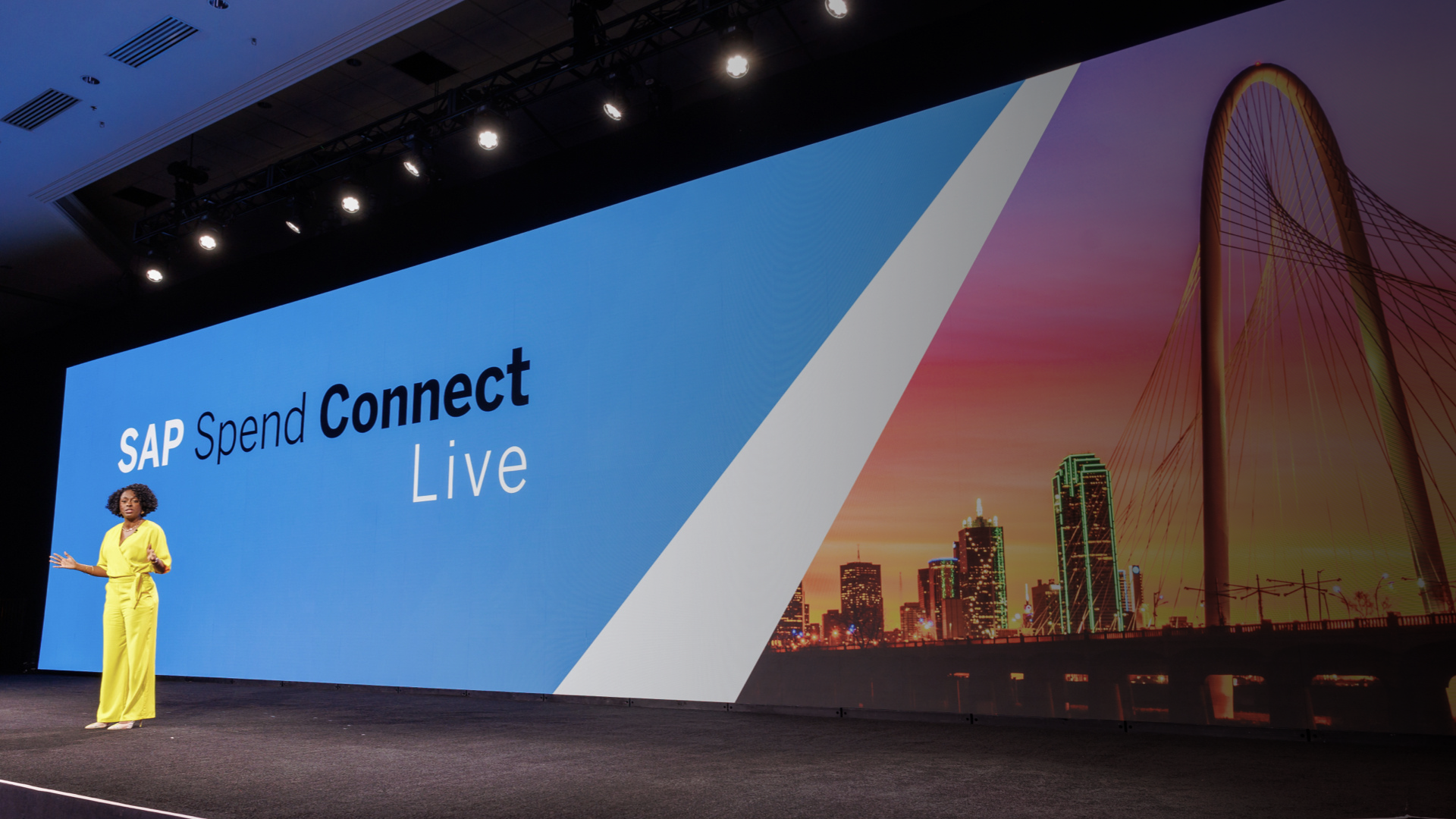 Photo of a woman giving a presentation in front of a blue screen that reads "SAP Spend Connect Live"