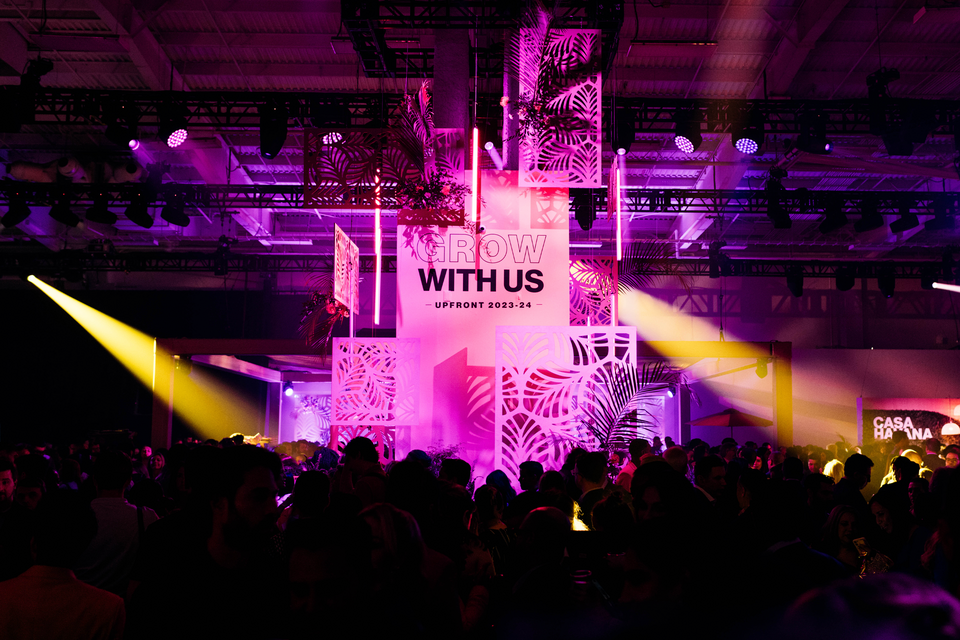 Photo of crowd looking at centerpiece reading Grow with Us Upfront 2023-2024. Scene has pink lighting