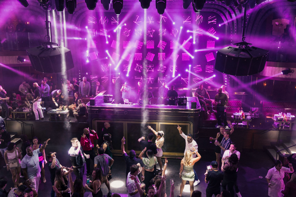 Photo inside a nightclub with a DJ, Magenta lighting, and crowd members dancing